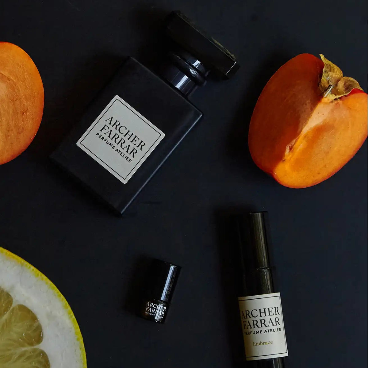 A dark matte perfume bottle and a matching roll-on bottle with cream labels from Archier Farrar, arranged on a dark surface with citrus slices and persimmon halves around them.