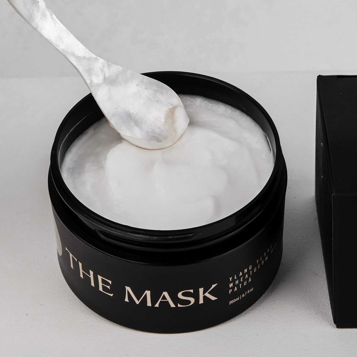 Black container of 'The Mask' with a spoonful of white mask on a light gray background
