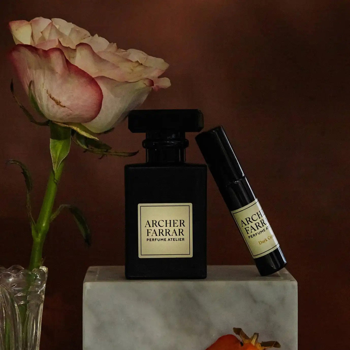 Two bottles of Archer Farrar perfume on a marble block with a rose and persimmon fruit.