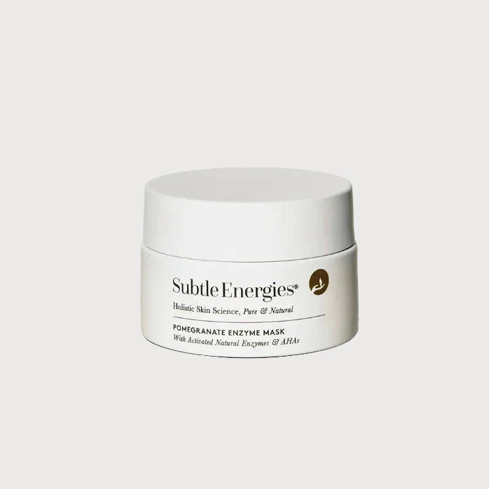 A white cosmetic jar of Subtle Energies Pomegranate Enzyme Mask with black text and a small gold circular icon, set on a light beige surface against a white background.