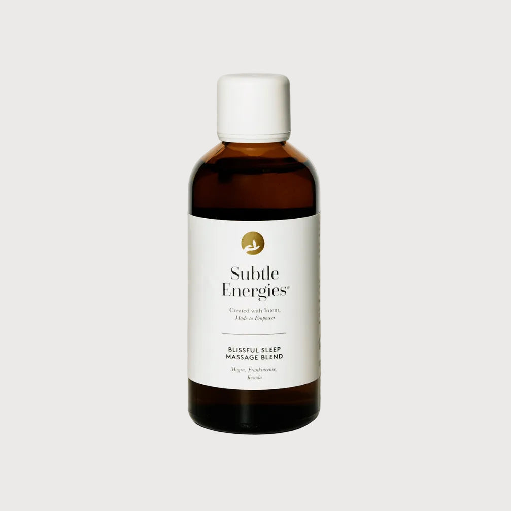 A bottle of Subtle Energies Blissful Sleep Massage Blend on a light beige surface against a white background. The bottle is made of dark amber glass with a white cap and a white label featuring a gold logo.