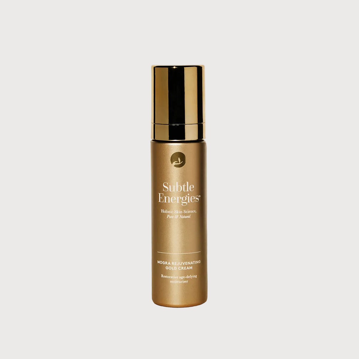 A gold cosmetic bottle labeled 'Subtle Energies' with a matching gold cap, standing on a light stone surface against a white wall with a soft shadow.