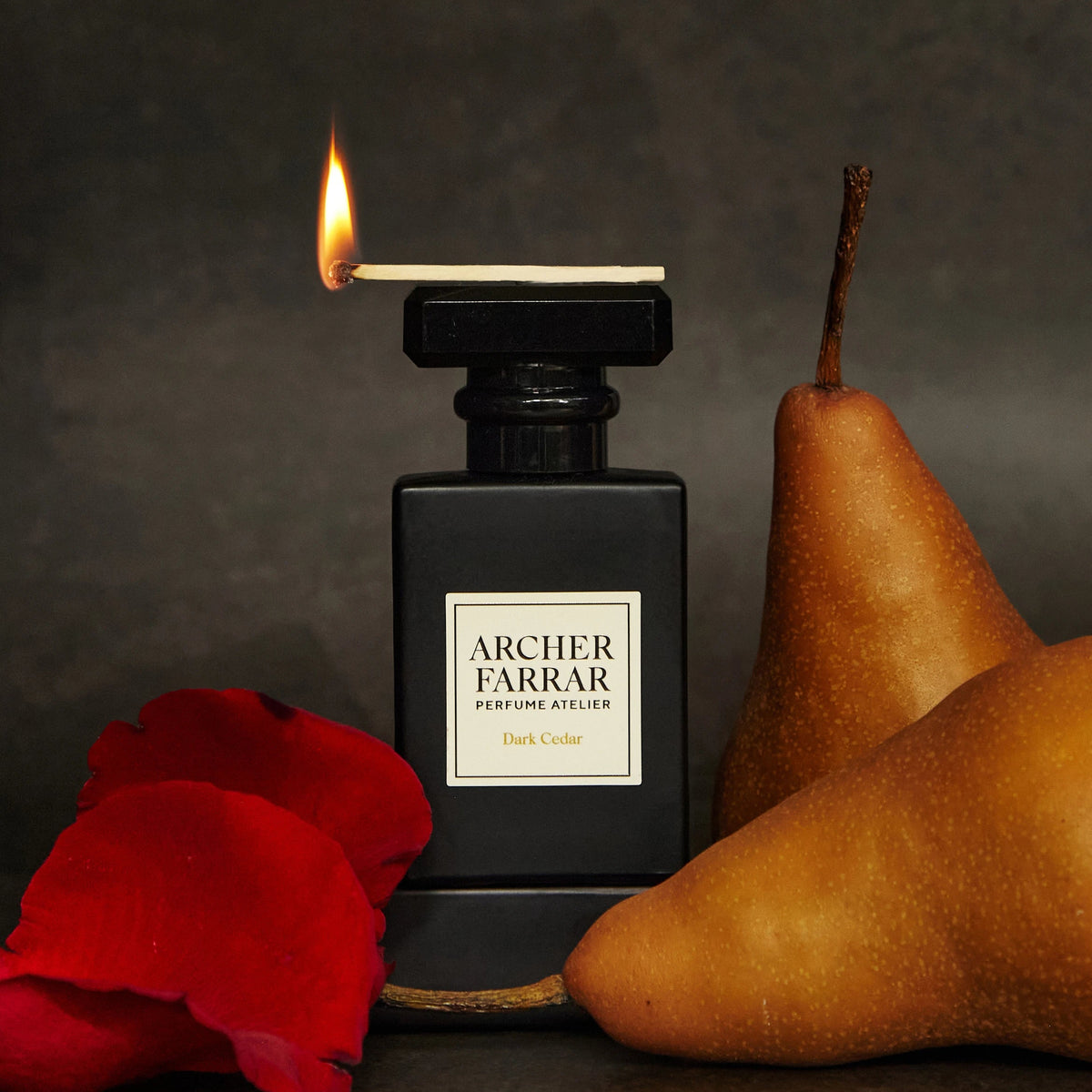 Black Archer Farrar perfume bottle with a lit match, red rose, and two pears on a dark background