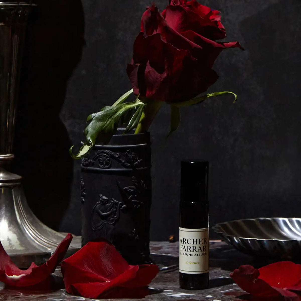 Dark room with a red rose in a vase and a bottle of Archers of Barakah perfume on a marble surface.