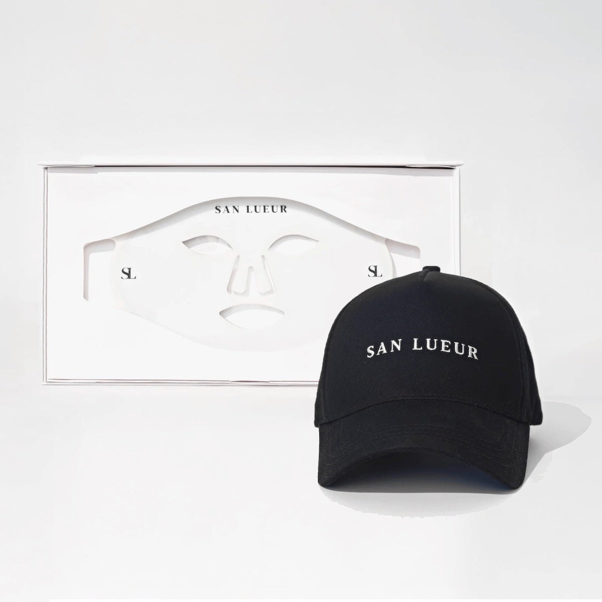 A black baseball cap with 'SAN LUEUR' embroidered in white on the front, displayed next to its white packaging box containing a matching white face mask on a clean white background.